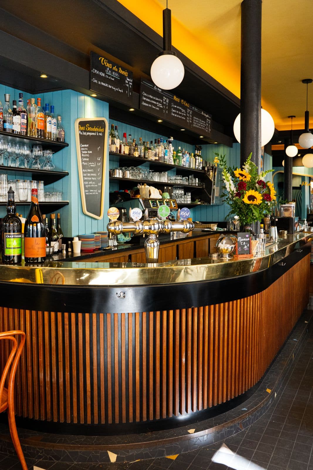 Le bar incurvé en zinc et le mur sarcelle du Roi de Pique, fleurs de tournesol sur le comptoir